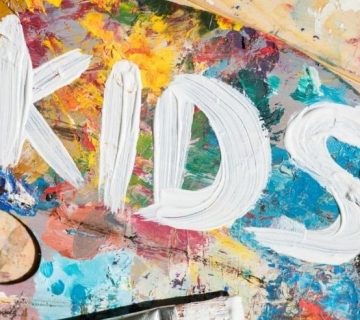 White paint spells out "KIDS" on a messy, colorful painter’s palette covered with splashes and strokes of various bright colors 
