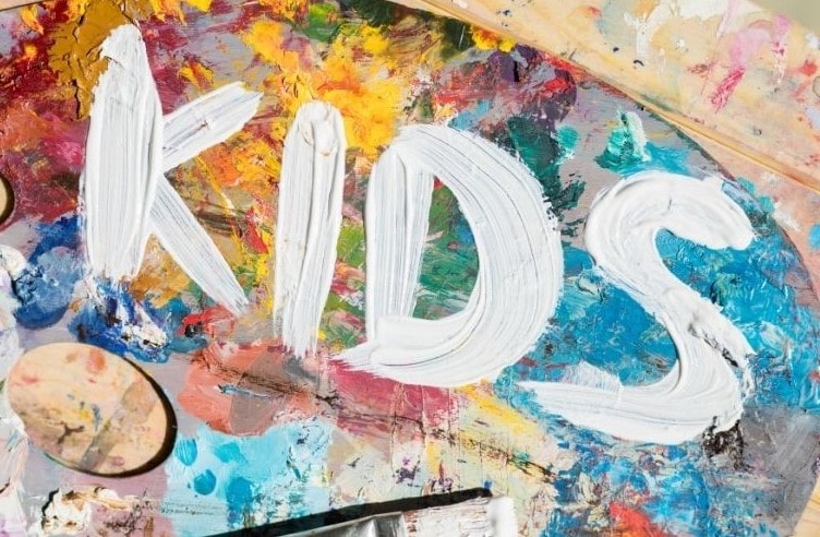 White paint spells out "KIDS" on a messy, colorful painter’s palette covered with splashes and strokes of various bright colors