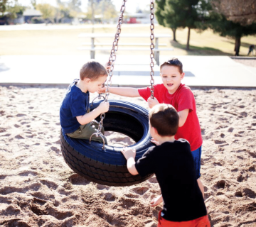 Three young boys play together on a black tire swing suspended by chains at a sandy playground, with trees and picnic tables visible in the background on a sunny day 
