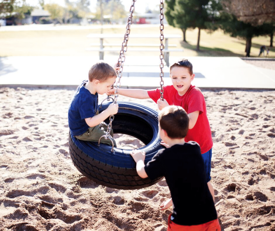 Three young boys play together on a black tire swing suspended by chains at a sandy playground, with trees and picnic tables visible in the background on a sunny day