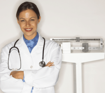 A smiling doctor in a white coat and stethoscope stands with arms crossed in front of a medical scale 