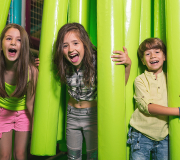 Three smiling children stand among tall, bright green soft foam tubes at an indoor play area, laughing and looking at the camera 