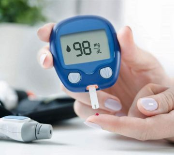 A person checks their blood sugar using a glucometer, which displays a reading of 98 mg/dL  One hand holds the meter; the other hand has a small drop of blood on a fingertip 