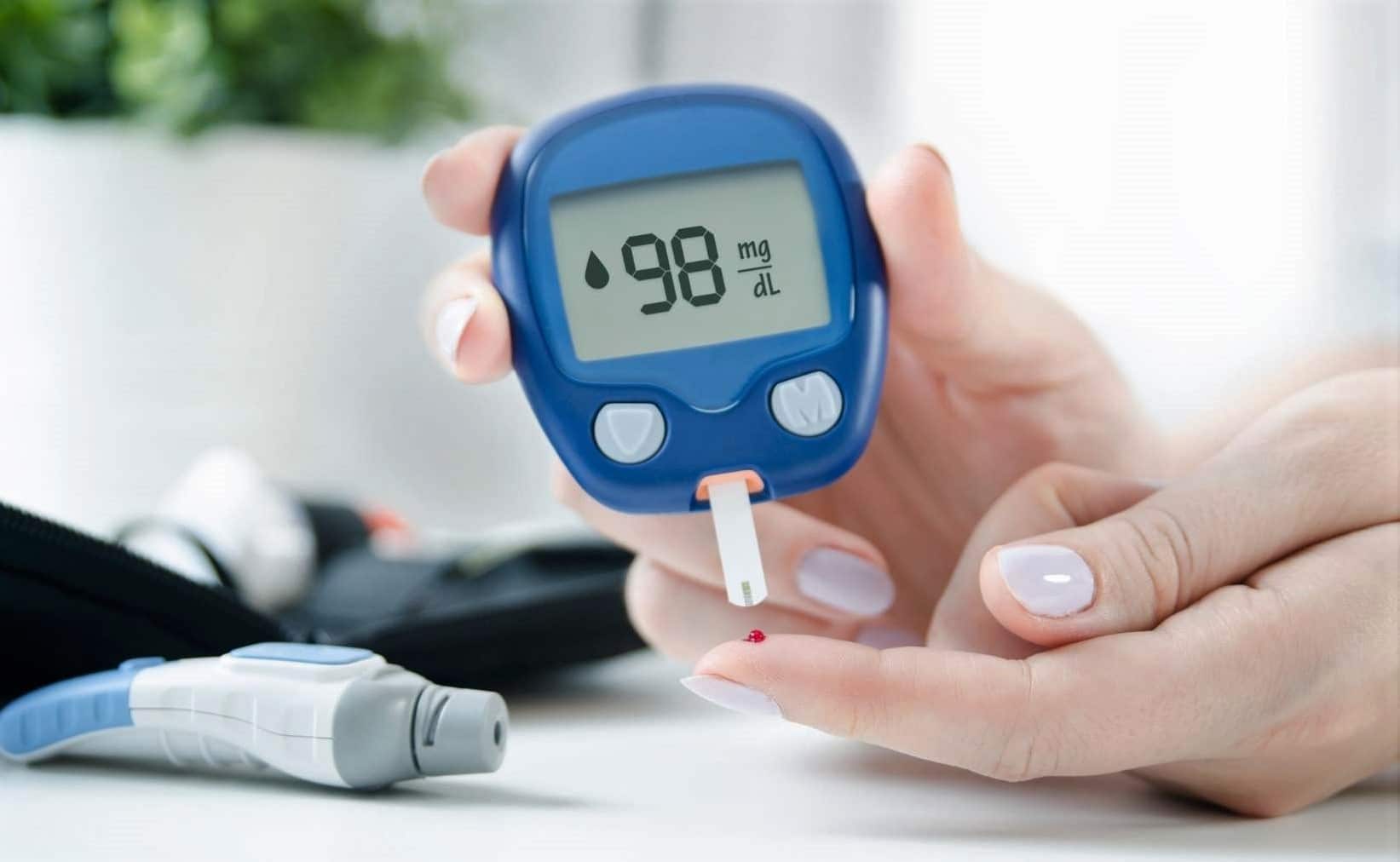 A person checks their blood sugar using a glucometer, which displays a reading of 98 mg/dL  One hand holds the meter; the other hand has a small drop of blood on a fingertip 